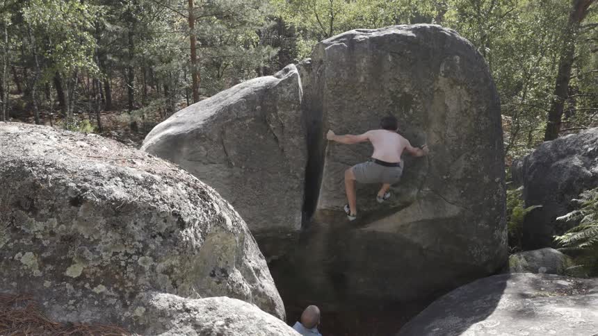 A person climbing the side of a large fissured rock formation in a wooded area while another person watches below