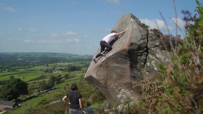 A man climbs a boulder as another watches in the countryside