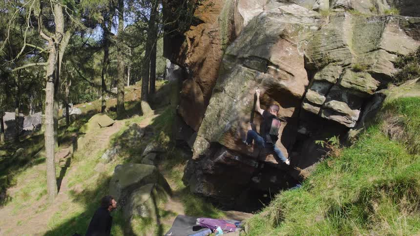 A man free climbs a large rock in the woods at midday while his friend watches nearby