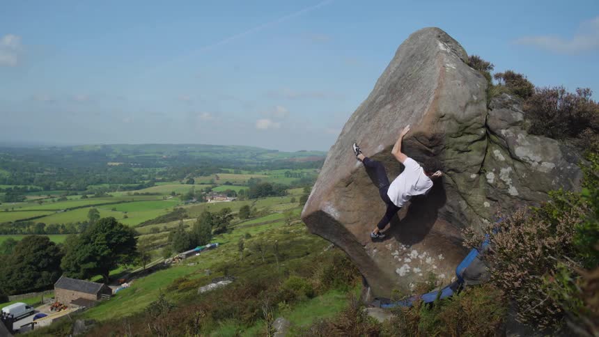 A man climbs a boulder on a hillside with farmland in the background