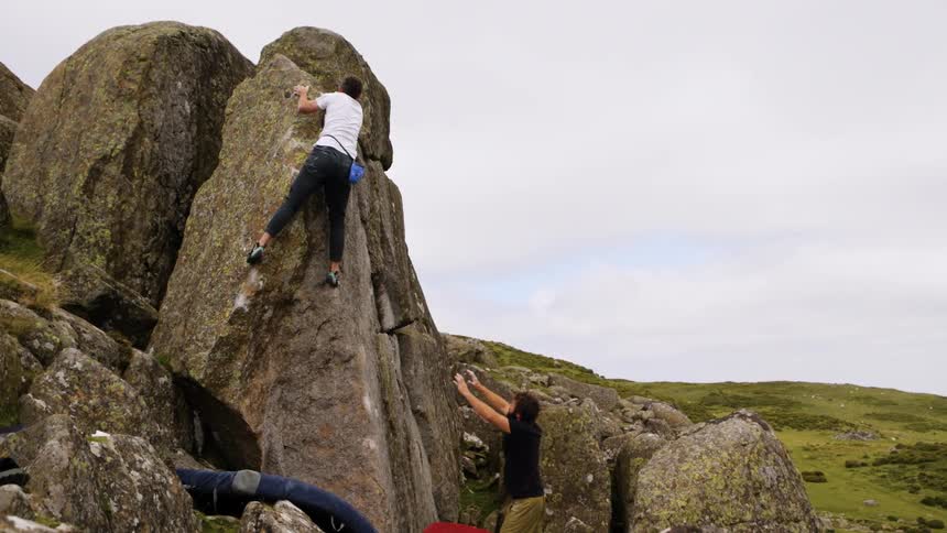 A man climbs a boulder as another spots him in a rocky landscape