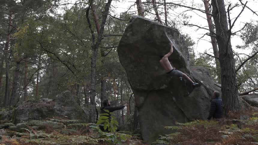 Man boulders on a large rock with two spotters nearby in a forest