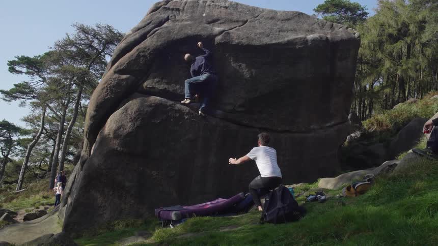 A young man climbing a large rock with the guidance of friends outdoors during daytime