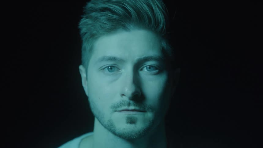 A closeup portrait of a young man with styled dark hair and a short beard transitions between vibrant colored lighting changing from purple to blue to green against a solid black background