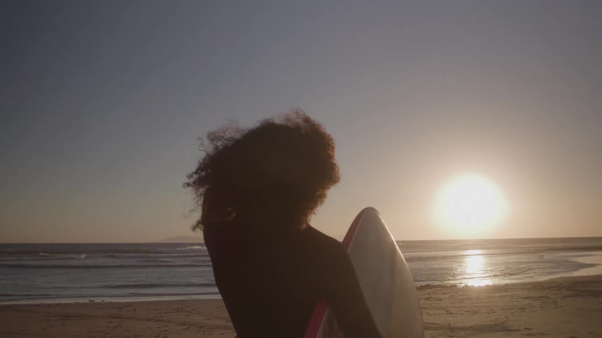 Woman shades her eyes from the sun while standing on the beach with a body board at sunset