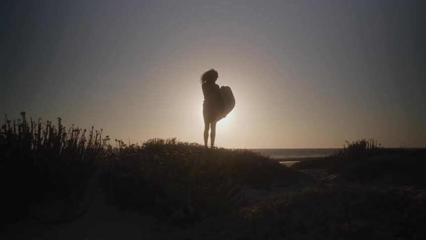 A woman stands on the shoreline at sunset holding a surfboard by her side