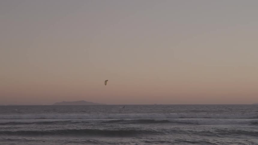 A lone kiteboarder is seen as a small silhouette on the choppy ocean waves with a single island visible on the distant horizon under an overcast or hazy evening sky