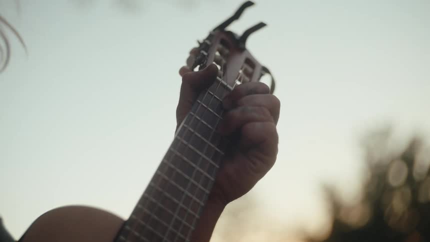 Closeup hands strum a small acoustic guitar at dusk warm bokeh framing the scene