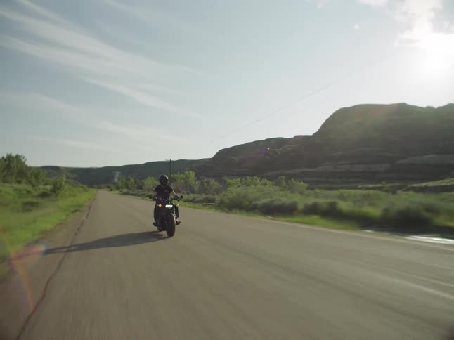 A person wearing dark clothes and a helmet rides a black motorcycle down a paved road that runs between grassy hills and utility poles casting a long shadow under a bright hazy sun
