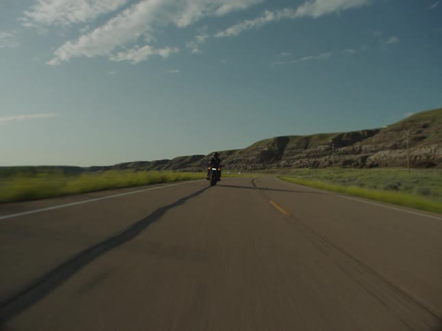 A person wearing dark clothes and a helmet rides a motorcycle directly toward the camera on an asphalt road bordered by green fields and arid hills under a partly cloudy blue sky