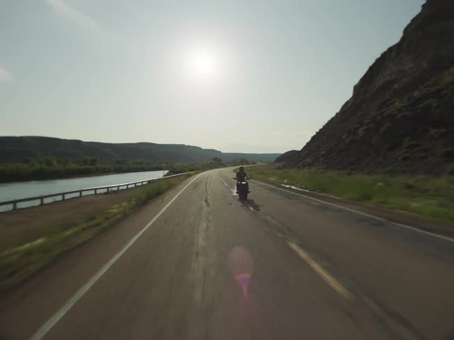 Motorcyclist rides along a road beside a river and cliff under the intense sun