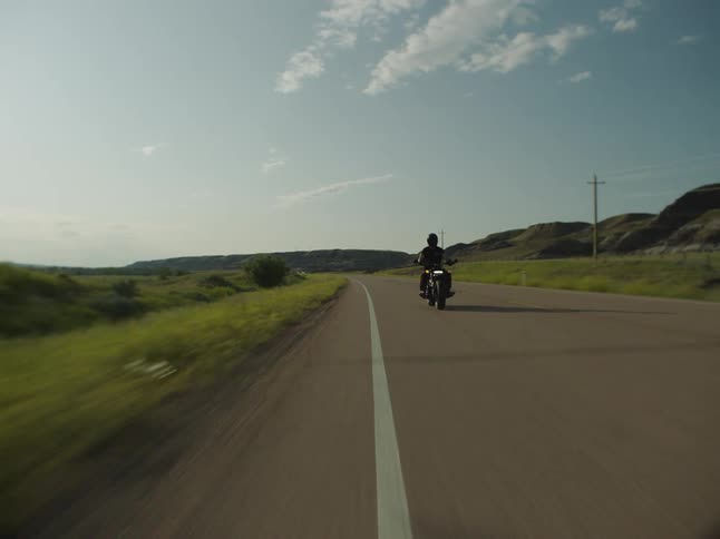 A motorcycle driving down a long straight country road surrounded by green hills under a bright sky