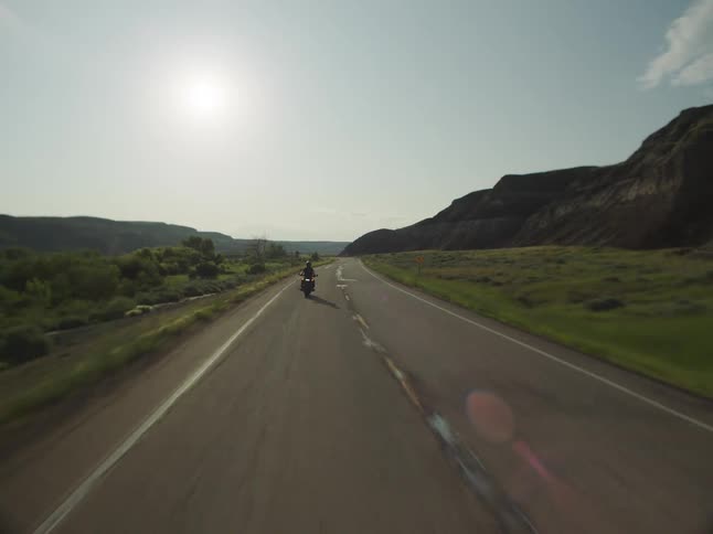 A person riding a motorcycle drives away from the camera down a paved road that extends into a bright sunlit valley with a large dark hill on the right and green low terrain on the left all under a mostly clear sky