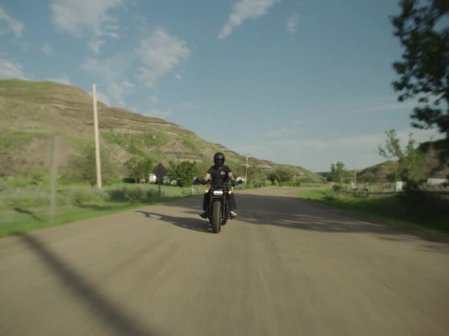 A person wearing black clothes and a helmet rides a dark motorcycle down a paved rural road crossing a wooden bridge with teal trusses surrounded by grassy hills on a sunny day