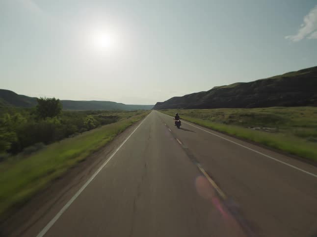 Motorcyclist rides along a long road surrounded by hills under the afternoon sun