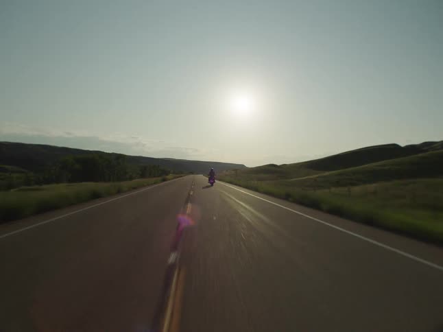 A motorcycle driving away on a long straight country road with the sun low in the sky causing a lens flare