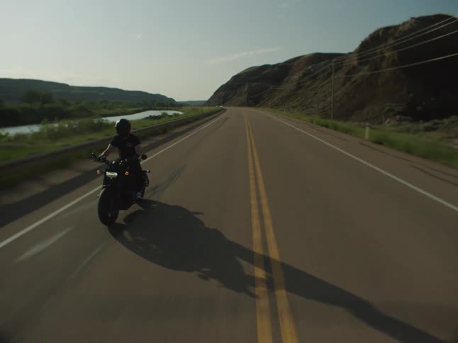 A person wearing a helmet and dark clothes rides a black motorcycle toward the camera on an asphalt road alongside a river and arid hills under the setting sun casting a long shadow