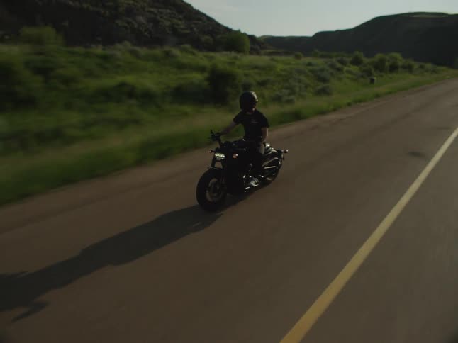 A biker on a motorcycle rides toward the camera along an asphalt road that cuts through green hills under a bright sun