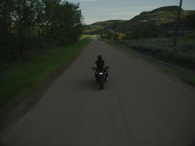 A person riding a dark motorcycle is filmed from behind as they ride down a paved road cross a turquoise metal and wooden bridge and continue down the road