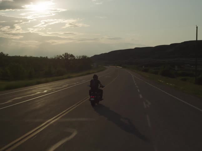 A person wearing a helmet and dark clothes rides a motorcycle away from the camera on an asphalt road bordered by arid hills driving directly toward the bright hazy afternoon sun