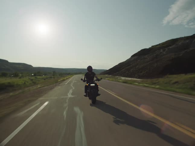 A person riding a motorcycle on a rural road passing hills and grass fields