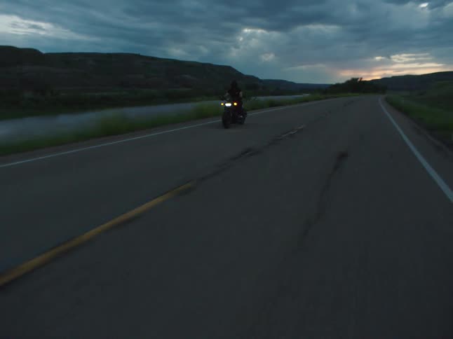 Motorcyclist rides along a road beside a river under the darkening dusk sky