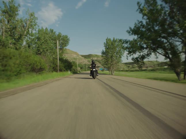 A biker rides a motorcycle down a scenic country road approaching and then crossing a green metal bridge in a wide sweeping shot