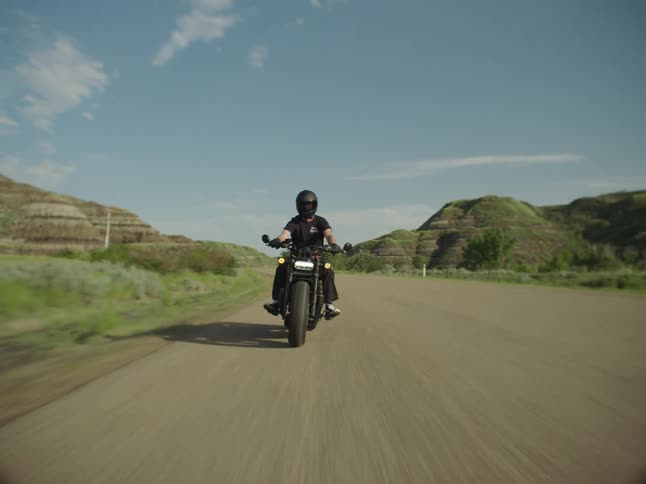 A person wearing a black helmet and dark shirt rides a black motorcycle directly toward the camera on an asphalt road bordered by arid green hills under a partly cloudy blue sky on a sunny day