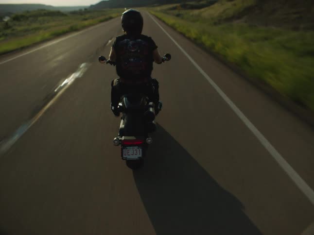 A man wearing a dark helmet and a backpack rides a black motorcycle straight down a sunny highway driving away from the camera through a green rolling landscape at sunset or sunrise