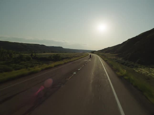 A person on a motorcycle rides along a straight empty highway framed by dark grassy hills driving away from the camera toward the bright setting sun that creates a strong lens flare