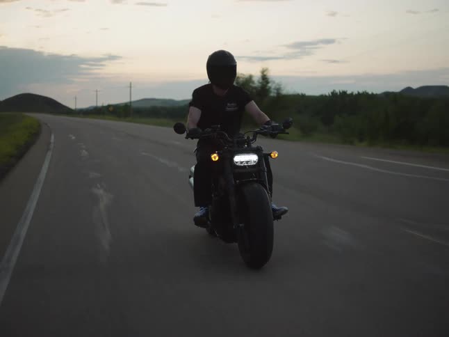A biker in a dark helmet and shirt rides a motorcycle along a country road at dusk or dawn with its headlights brightly lit