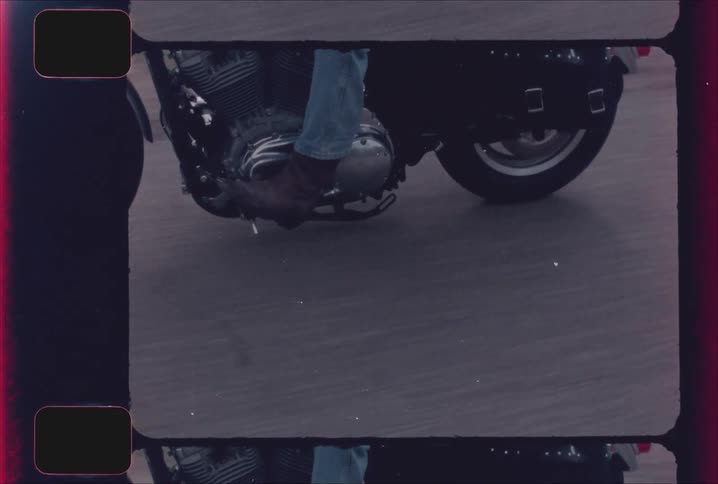 A low close view shows the engine and front wheel of a cruiser motorcycle with a riders leg in jeans and a brown boot shot with a vintage film effect that includes sprocket holes and film markings on the edge