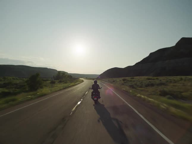 A silhouette of a person on a motorcycle rides along a straight highway road with white road markings driving directly toward the bright hazy setting sun between dark arid hills