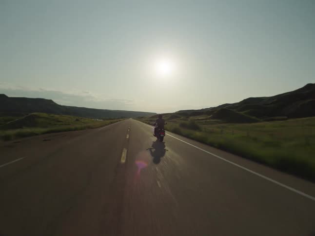 A lone motorcyclist cruises a sunlit twolane highway through rolling green hills and open country