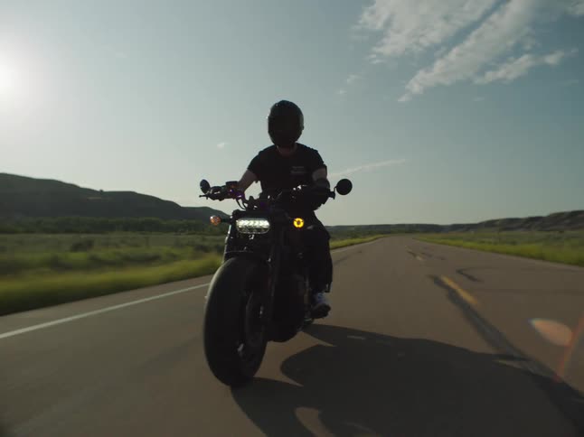 Male motorcyclist rides on a rural highway passing through the fields between hills on a sunny day