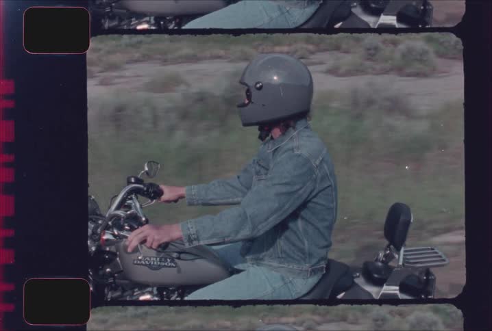 A person in a grey helmet sunglasses and denim jacket riding a harley davidson motorcycle