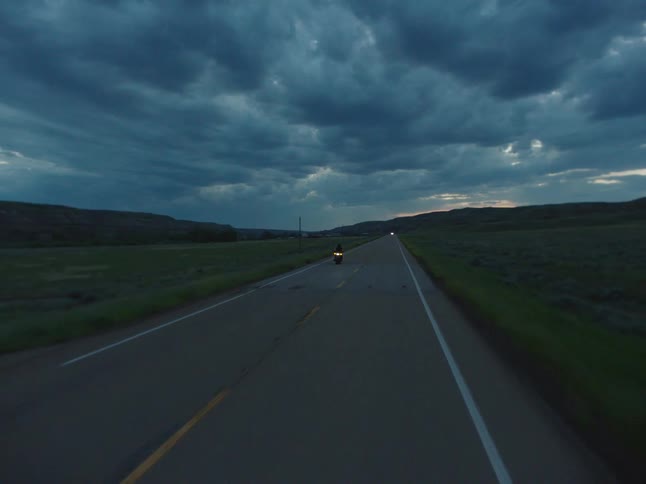 A person on a motorcycle rides on a rural road across a long grassy plain under a cloudy sky