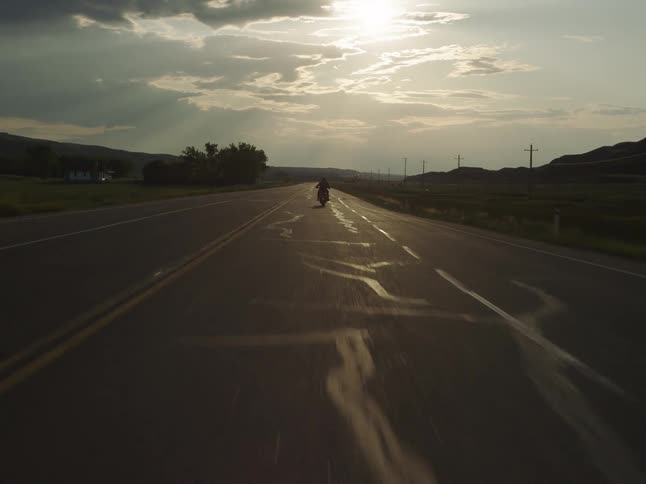 Motorcyclist rides along the road at sunset with the sun among clouds