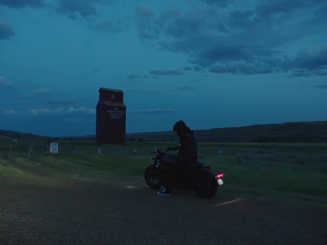 A person on a motorcycle parked by the side of a rural road at night next to a tall barn in a grass field