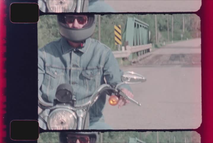 A man wearing a jacket helmet and sunglasses rides a motorcycle along a countryside road on a sunny day