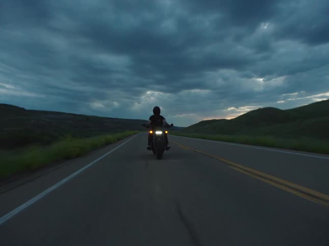 A person in a helmet rides a motorcycle with headlights on along a dark empty rural road moving away from the camera into a valley between shadowed hills under a dramatic dark cloudy sky