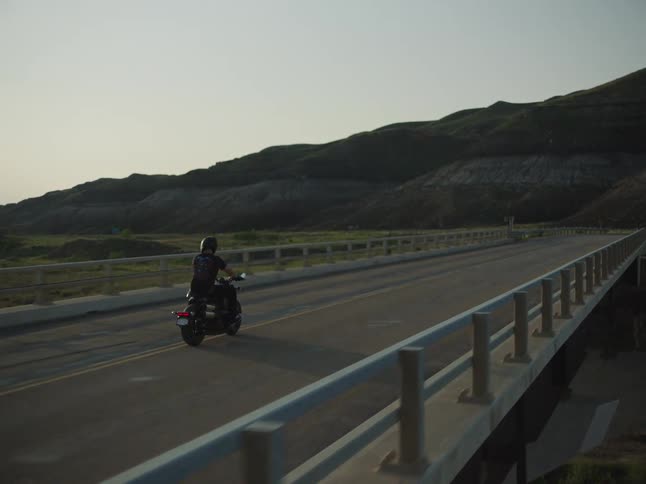 A biker on a motorcycle rides across a bridge on a winding road disappearing into the vast hilly and dramatic landscape