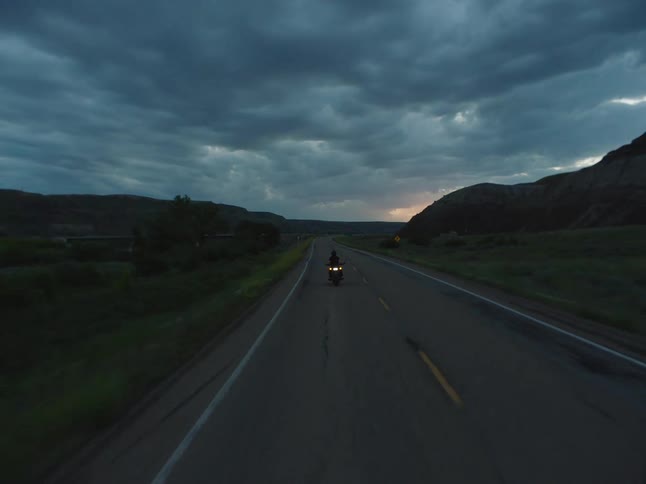 A silhouette of a person on a motorcycle drives along a dark straight highway surrounded by arid hills driving toward the fading light of sunset beneath a dramatic cloudy twilight sky
