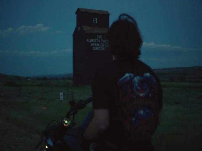 A man wearing a dark helmet and a graphic tshirt sits on a motorcycle with the headlight on looking at a dark tall grain elevator in a field beneath a cloudy twilight sky