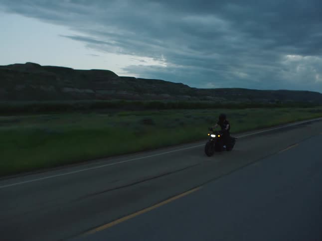 Motorcyclist rides along a deserted road at dusk with hills in the background