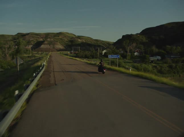 Motorcyclist rides along a road surrounded by green hills at sunset