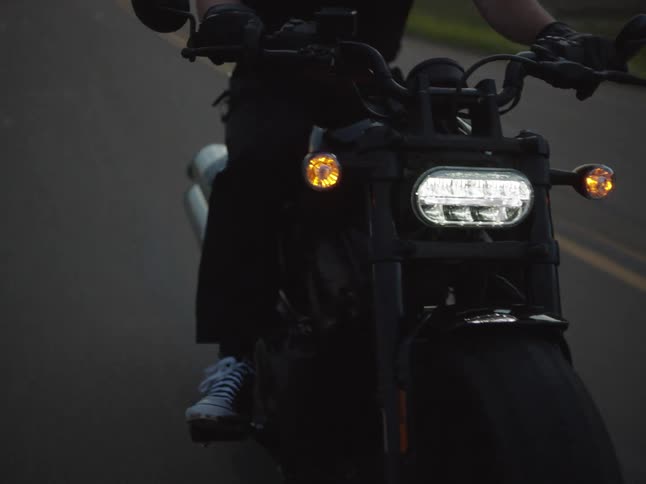 A man wearing a dark helmet and black tshirt rides a black motorcycle with its bright headlight on directly toward the camera on a rural road in a hilly landscape at dusk