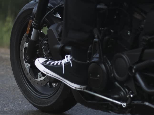 A person wearing black sneakers rides a motorcycle across the road at midday