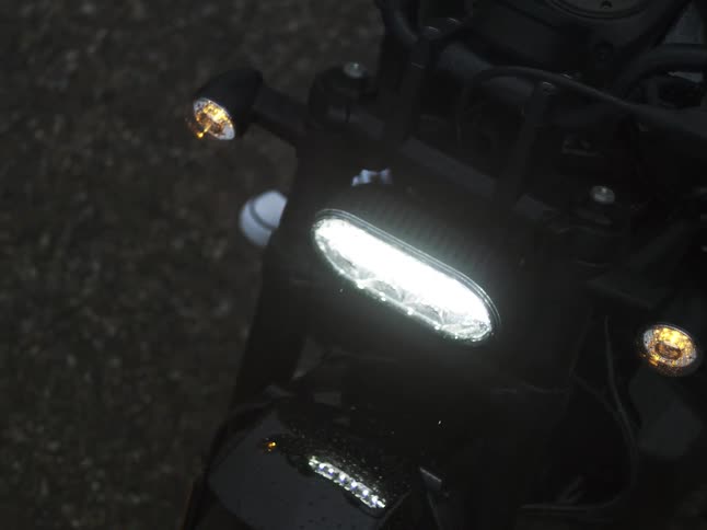 A close overhead view shows a gloved hand pressing the ignition button on a dark motorcycle causing the bright led headlight and orange turn signals to flash and illuminate the wet asphalt in the dark
