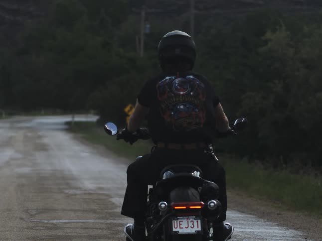 A man wearing a helmet and a dark tshirt with a graphic on the back rides a black motorcycle away from the camera on a dark paved rural road surrounded by shadowed trees at dusk